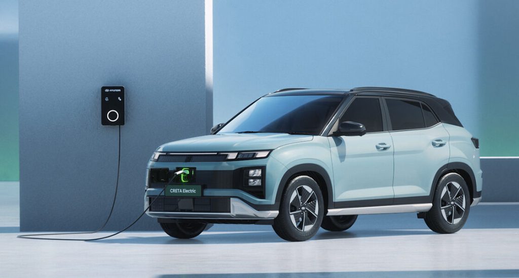 Front three-quarter view of a light-blue Hyundai Creta Electric SUV plugged into a Hyundai wall-mounted home charging station against a minimalist backdrop